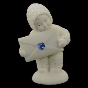 Puede incluir: Una figurita de porcelana blanca de un muñeco de nieve que lleva un sombrero blanco y sostiene un sobre blanco con una gema azul en forma de corazón.