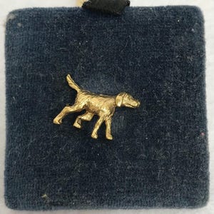 May include: A gold-toned metal pin shaped like a dog in a running stance, featuring a textured surface and detailed limbs. The pin is attached to a square, dark blue, velvet-covered display pad with a small black fabric tab at the top edge.