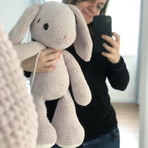 May include: A light gray crocheted bunny plush toy with long floppy ears, black eyes, and a pink nose. The handmade stuffed animal has a textured surface and is being held up in front of a mirror.