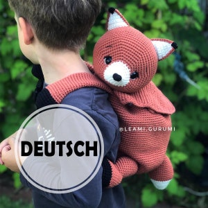 Peut inclure: Un enfant portant un sac à dos en crochet en forme de renard. Le sac à dos est brun avec des accents blancs et des finitions noires. Le texte "DEUTSCH" est visible sur le sac à dos.