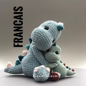May include: Two crocheted plush dinosaurs, one blue and one green, are hugging. The larger blue dinosaur has pink accents on its feet. The smaller green dinosaur has black eyes. The dinosaurs are sitting on a grey surface.