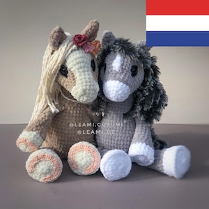 Op de afbeelding: Twee handgemaakte gehaakte paardenknuffels. Eén is bruin met een crème manen en een bloemenkroon. De andere is grijs met een wit gezicht en een pluizige manen. Beide hebben witte hoeven en zitten naast drie ronde, gehaakte ballen.