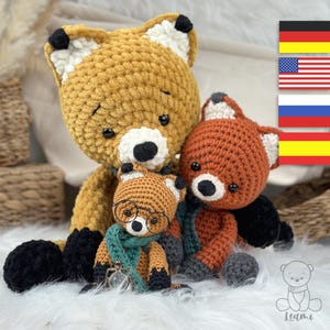 May include: Three crocheted fox plushies, one large, one medium, and one small. The large fox is mustard yellow, the medium fox is orange, and the small fox is orange with green glasses and a green scarf. The plushies are sitting on a white fluffy surface.
