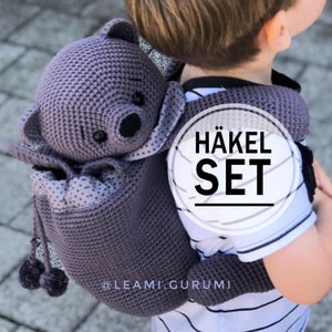 Könnte beinhalten: Ein lilafarbener Häkel-Bärenrucksack mit Kordelzug und einem passenden Paar Häkel-Handschuhen. Der Rucksack hat ein gepunktetes Futter und der Text "HÄKEL SET" ist auf der Vorderseite sichtbar. Der Rucksack wird von einem Kind getragen.
