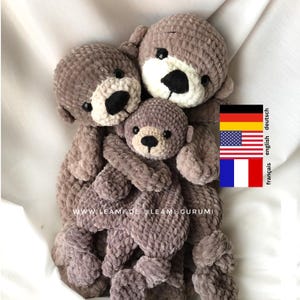 May include: Three crocheted plush otters in a hug. The otters are brown and have black eyes and noses. The otters are sitting on a white surface. The text "www.leamiguru.com" is visible on the bottom of the image.