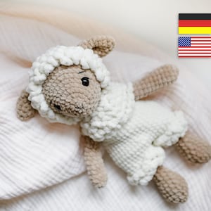 Könnte beinhalten: Ein weißes, gehäkeltes Lamm-Plüschtier mit braunen Augen und einer braunen Nase. Das Spielzeug liegt auf einer weißen Decke.