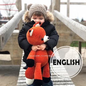 Könnte beinhalten: Ein Kind in einer schwarzen Jacke und einer braunen Strickmütze hält ein gehäkeltes orangefarbenes Fuchs-Plüschtier. Das Spielzeug hat schwarze Augen, eine weiße Nase und schwarze und weiße Akzente an seinen Beinen und seinem Schwanz. Das Wort "ENGLISH" ist in weißen Buchstaben auf einem schwarzen Kreis geschrieben.