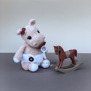 Può includere: Un ippopotamo di peluche all'uncinetto che indossa un pannolino bianco e un ciuccio rosa, seduto accanto a un cavallo a dondolo di legno. L'ippopotamo ha un fiocco rosa sulla testa e occhi neri. Il cavallo a dondolo è marrone con una sella bianca.