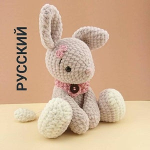 May include: A crocheted plush bunny toy with a pink bow, a pink collar with a black button, and white feet. The bunny is sitting on a light brown surface.