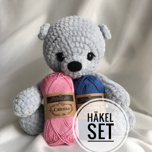 Könnte beinhalten: Ein hellblauer gehäkelter Teddybär mit schwarzen Augen und einer schwarzen Nase. Der Bär hält zwei Knäuel Garn, eines rosa und eines blau, mit dem Text "Scheepjes Catona" auf den Etiketten. Ein weißer Kreis mit dem Text "HÄKEL SET" befindet sich im Bild.