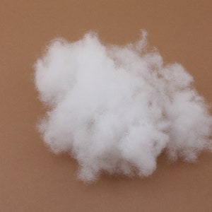 Könnte beinhalten: Ein Haufen weicher, weißer Polyester-Faserfüllung auf einem einfarbigen, braunen Hintergrund. Das Material, das zum Füllen von Kissen und Spielzeug verwendet wird, hat ein wolkenartiges Aussehen. Das Bild ist eine Nahaufnahme, die die Textur zeigt.