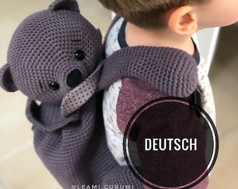 Pode incluir: Uma mochila de urso de peluche de crochê cinza com olhos pretos e nariz preto. A mochila está sendo usada por uma criança. O texto "DEUTSCH" é visível em um círculo na mochila.