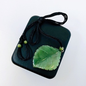 Puede incluir: Un collar con un colgante de hoja de jade verde, un cordón negro y cuentas verdes. El collar se muestra en una caja de terciopelo negro.