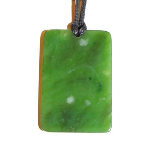 May include: A green jade pendant with a dark grey cord. The pendant is rectangular and has a smooth, polished surface.