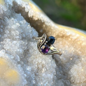 May include: A silver ring featuring a black and purple gemstone setting. The ring is displayed against a white, crystalline background.