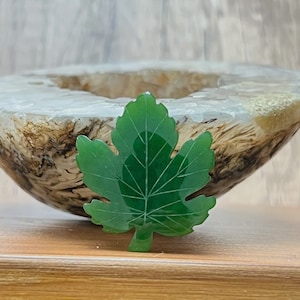 Könnte beinhalten: Ein grüner Jade-Anhänger in Blattform mit einem detaillierten Adernmuster. Der Anhänger liegt auf einer Holzoberfläche.