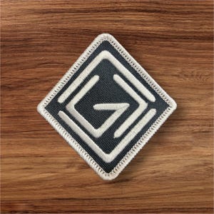 May include: A square patch with a dark gray center and a white geometric design. The design is a stylized letter "G" within a diamond shape, outlined with white stitching. The patch is on a wooden surface.