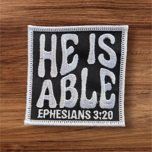May include: A square black patch with white embroidered text that reads "HE IS ABLE" and "EPHESIANS 3:20". The patch has a white border and is set against a wooden background.