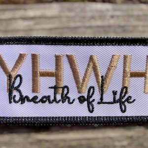 May include: A rectangular embroidered patch with a white background and a thick black border. The letters YHWH are stitched in metallic gold thread, while the phrase Breath of Life is embroidered in black cursive script underneath the letters.
