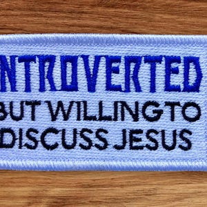 May include: A light blue embroidered patch with the text "Introverted but willing to discuss Jesus" in black and blue thread.