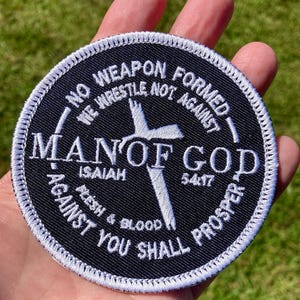 May include: A round, black patch with white embroidered text and a white cross design. The text includes "NO WEAPON FORMED," "MAN OF GOD," and biblical verses. The patch has a white stitched border.