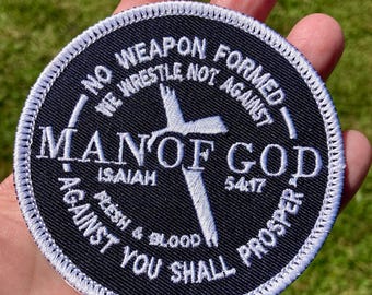 May include: A round, black patch with white embroidered text and a white cross design. The text includes "NO WEAPON FORMED," "MAN OF GOD," and biblical verses. The patch has a white stitched border.