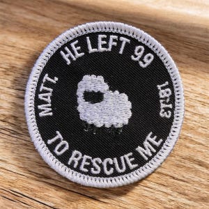 May include: Round black patch with white embroidered text and a sheep illustration. The text reads "He Left 99 To Rescue Me" and "Matt 18:13". The patch has a white stitched border and is set against a wooden surface.