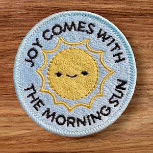 May include: Embroidered patch with a light blue background and the text "JOY COMES WITH THE MORNING SUN" in black. The patch features a smiling yellow sun in the center, with a gold outline. The patch is round and has a light blue border.