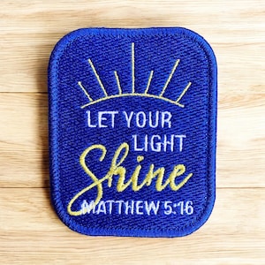 May include: A blue embroidered patch with the words "LET YOUR LIGHT Shine" in white and yellow script. The patch also includes the text "MATTHEW 5:16" and a sunburst design.