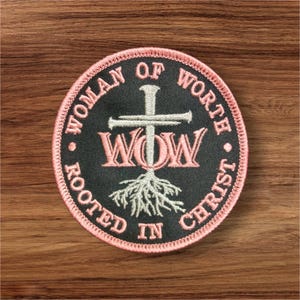 May include: Round embroidered patch with the text "WOMAN OF WORTH ROOTED IN CHRIST" in a circular pattern. The patch features a cross, the letters "WOW", and a root design in pink and white on a black background, with a pink border.