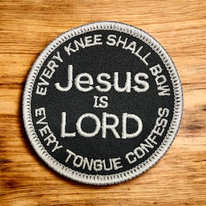 May include: A round black patch with white embroidered text. The text reads "Jesus IS LORD" in the center, surrounded by "EVERY KNEE SHALL BOW EVERY TONGUE CONFESS". The patch has a white stitched border.