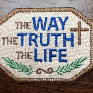 May include: An octagonal embroidered patch with a cream-colored background and a brown border. The text reads THE WAY THE TRUTH THE LIFE in blue and brown thread. A brown cross is on the right, and green leafy branches are at the bottom.