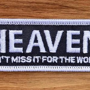 May include: A rectangular patch with the word "HEAVEN" in large white embroidered letters on a black background. Below, the text "DON'T MISS IT FOR THE WORLD" is also embroidered in white. The patch has a white border.