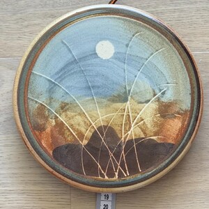 May include: Round ceramic plate with a landscape design. The plate features a blue sky with a white moon, brown and tan hills, and white grass-like lines. The rim is brown. The plate is approximately 9 inches in diameter.