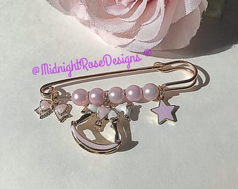 Può includere: Uno spillo di sicurezza dorato con perline rosa e bianche, un ciondolo a forma di cavallo a dondolo rosa e un ciondolo a forma di stella rosa. Lo spillo è su uno sfondo bianco.