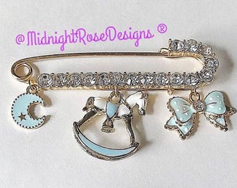 Può includere: Una spilla di sicurezza dorata con una fila di strass trasparenti. La spilla ha tre ciondoli: una mezzaluna blu, un cavallo a dondolo e un fiocco blu con pois bianchi. Il testo "@MidnightRoseDesigns®" è visibile nell'angolo in alto a sinistra dell'immagine.