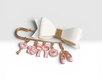 Può includere: Una spilla da balia color oro con un grande fiocco in tessuto bianco e un centro glitterato dorato. Dalla spilla pendono cinque ciondoli: una chiave rosa a forma di cuore, un fiore rosa con una perla, un fiocco rosa, una corona e scarpette da danza.