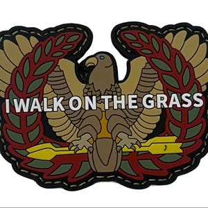 May include: A patch featuring an eagle with outstretched wings, surrounded by olive branches and a black border. The text "I WALK ON THE GRASS" is displayed in white letters across the center of the patch.