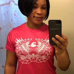 May include: A pink t-shirt with a white eagle graphic. The eagle is holding a rifle and is surrounded by a wreath of leaves. The shirt has short sleeves and a crew neck.