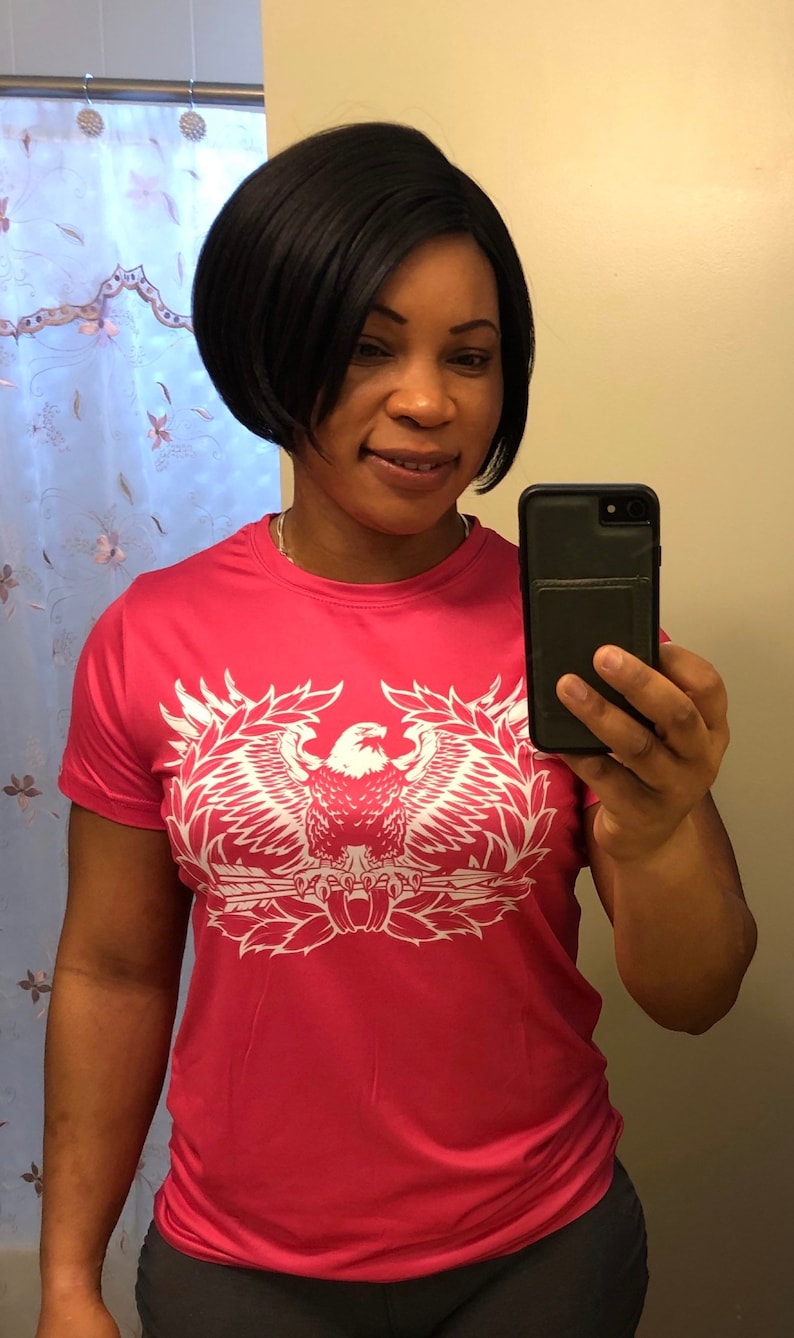 May include: A pink t-shirt with a white eagle graphic. The eagle is holding a rifle and is surrounded by a wreath of leaves. The shirt has short sleeves and a crew neck.