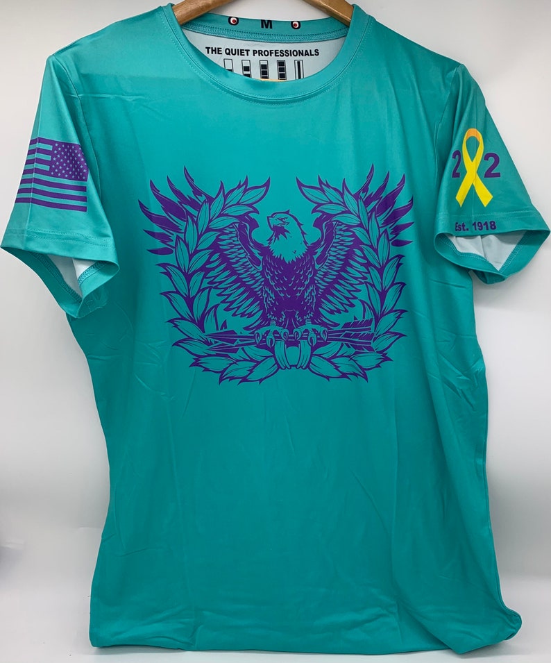 May include: Teal blue short-sleeved t-shirt with a purple eagle graphic and the text "The Quiet Professionals" on the front. The shirt also features a purple American flag on the left sleeve and a yellow ribbon with the number "22" and the text "Est. 1918" on the right sleeve.