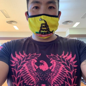 May include: A man wearing a black t-shirt with a pink eagle graphic and a yellow face mask with a snake and the text "Don't Cough On Me".