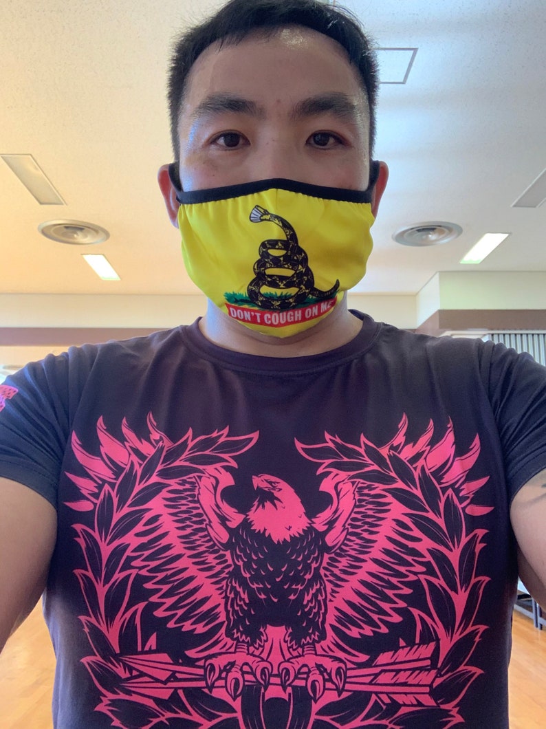 May include: A man wearing a black t-shirt with a pink eagle graphic and a yellow face mask with a snake and the text "Don't Cough On Me".