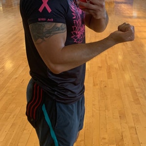May include: A man wearing a black t-shirt with a pink ribbon and a floral design, black shorts with red and blue stripes, and grey sneakers. He is wearing a yellow face mask and holding a red phone with a white case that says "2ubs" in white letters. The man is standing in a gym with a wooden floor.