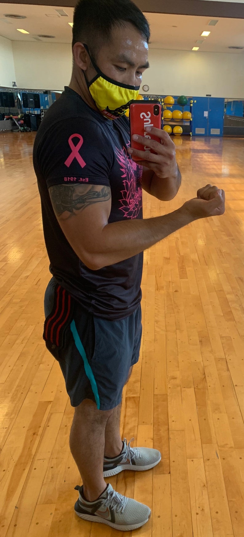May include: A man wearing a black t-shirt with a pink ribbon and a floral design, black shorts with red and blue stripes, and grey sneakers. He is wearing a yellow face mask and holding a red phone with a white case that says "2ubs" in white letters. The man is standing in a gym with a wooden floor.
