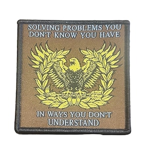 May include: A square patch with the text "SOLVING PROBLEMS YOU DON'T KNOW YOU HAVE IN WAYS YOU DON'T UNDERSTAND." The patch features a golden eagle with outstretched wings, surrounded by olive branches, on a brown background.