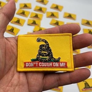 May include: Yellow rectangular patch featuring a coiled snake illustration with the text "DON'T COUGH ON ME" in red. The patch is held in a hand, with several similar patches visible in the background.