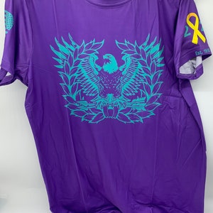 May include: Purple short-sleeved t-shirt with a teal eagle graphic and the text "The Quiet Professionals" on the front. The right sleeve has a yellow ribbon with the text "Est. 1918".