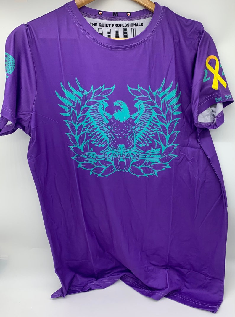 May include: Purple short-sleeved t-shirt with a teal eagle graphic and the text "The Quiet Professionals" on the front. The right sleeve has a yellow ribbon with the text "Est. 1918".