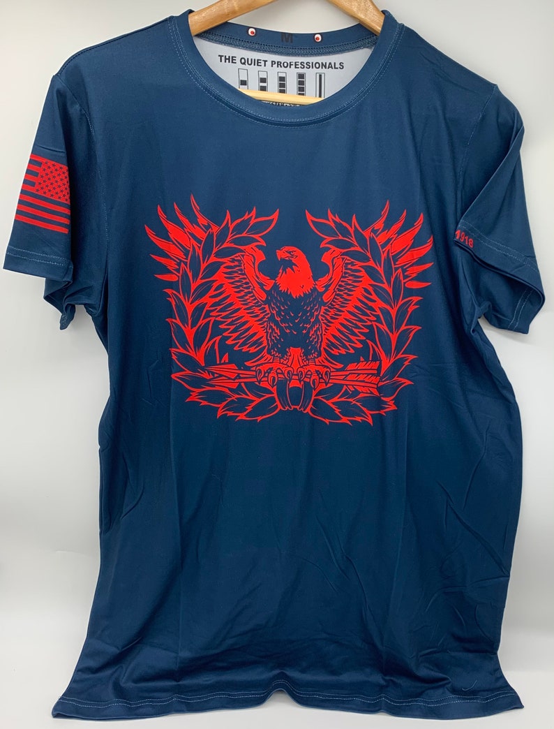 May include: A navy blue t-shirt with a red eagle graphic. The eagle is holding a rifle and is surrounded by a wreath. The text "THE QUIET PROFESSIONALS" is printed on the shirt.